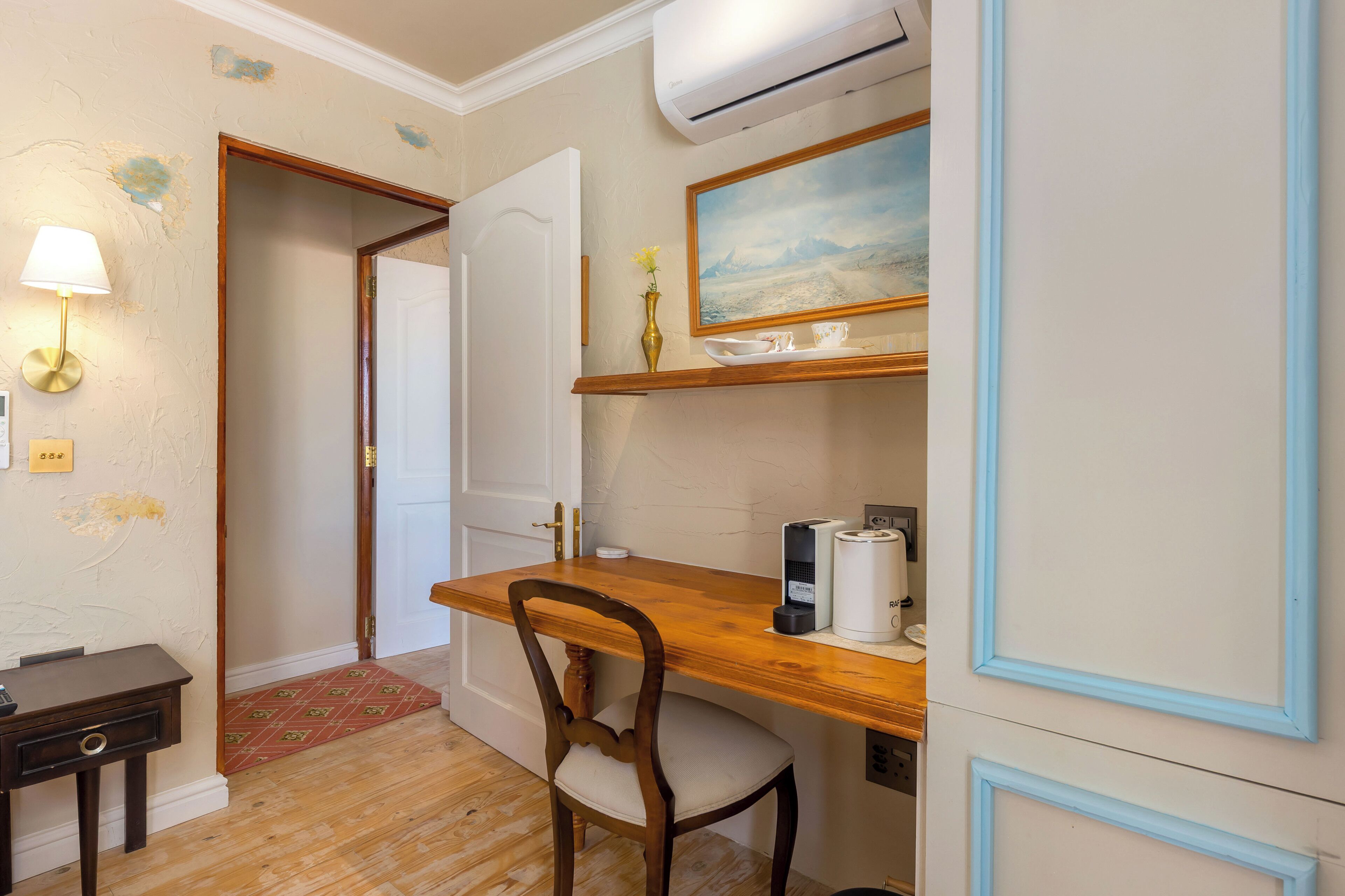 Una habitación con escritorio de madera, una silla, una mesita, aire acondicionado, un cuadro y una lámpara.