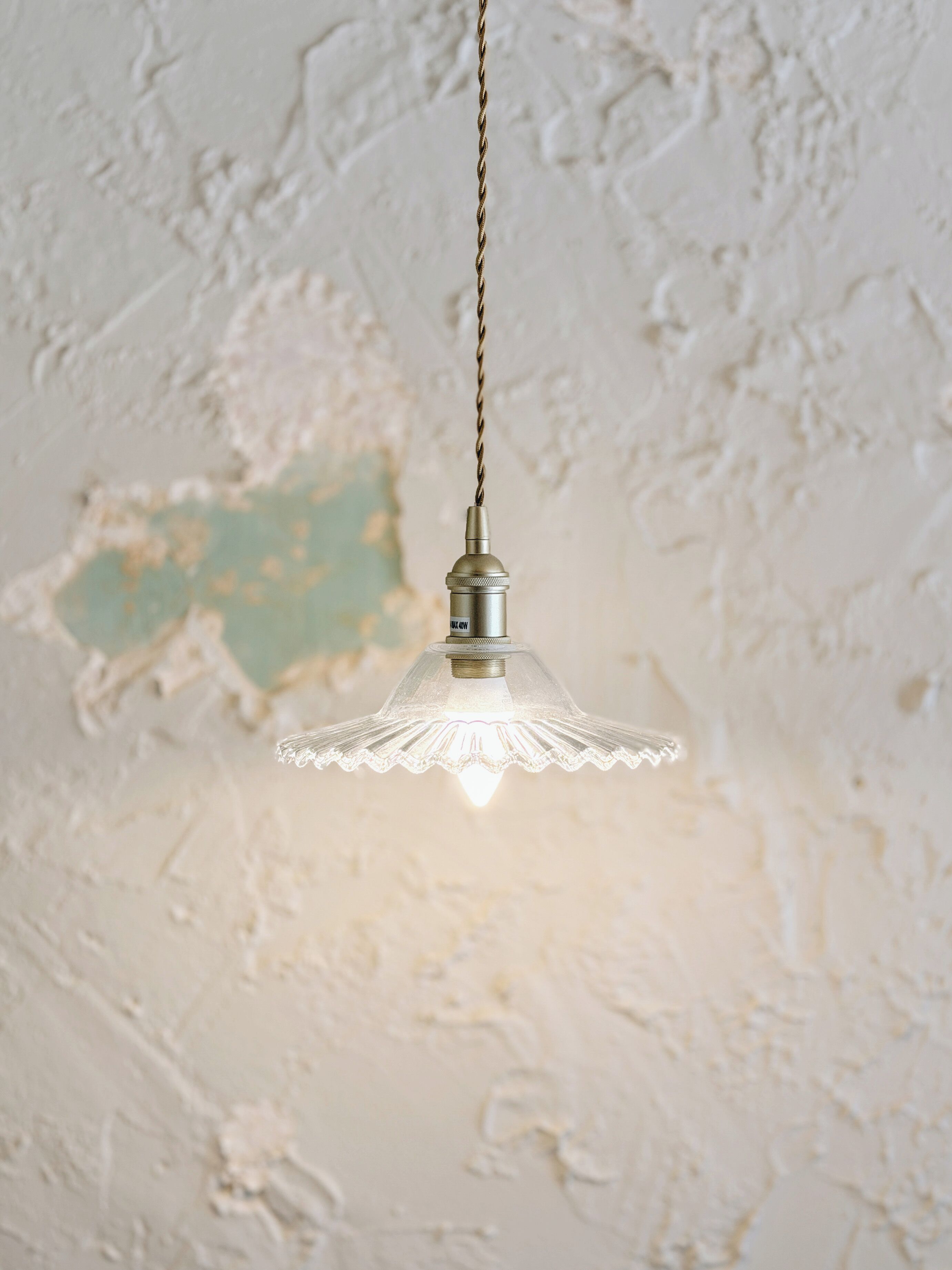 Lámpara colgante con pantalla de vidrio transparente y base de metal, suspendida de una cadena frente a una pared con textura.