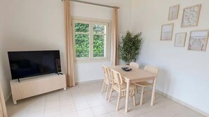 Un'ampia stanza con una televisione a schermo piatto, un tavolo da pranzo con sedie, una grande finestra con tende e un lampadario.