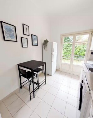Eine kleine Küche mit einem schwarzen Tisch und Stühlen, einem weißen Kühlschrank und einer Glastür, die auf einen Balkon führt.
