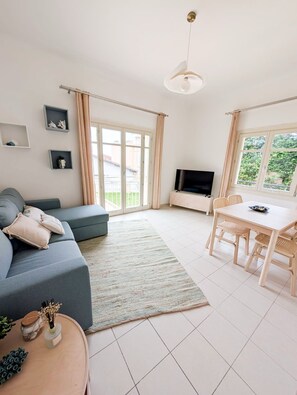 Ein modernes Wohnzimmer mit Sofa, Fernseher, Esstisch und Blick durch große Fenster.