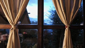 Ein Fenster mit zur Oberseite gebundenen Vorhängen, das einen Blick auf einen Garten mit Kübelpflanzen und einer Holzkonstruktion freigibt.