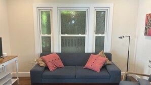 Ein Wohnzimmer mit einem dunkelblauen Sofa, zwei roten Kissen, einem Holztisch, einer Stehlampe und einem Fenster mit Milchglas.