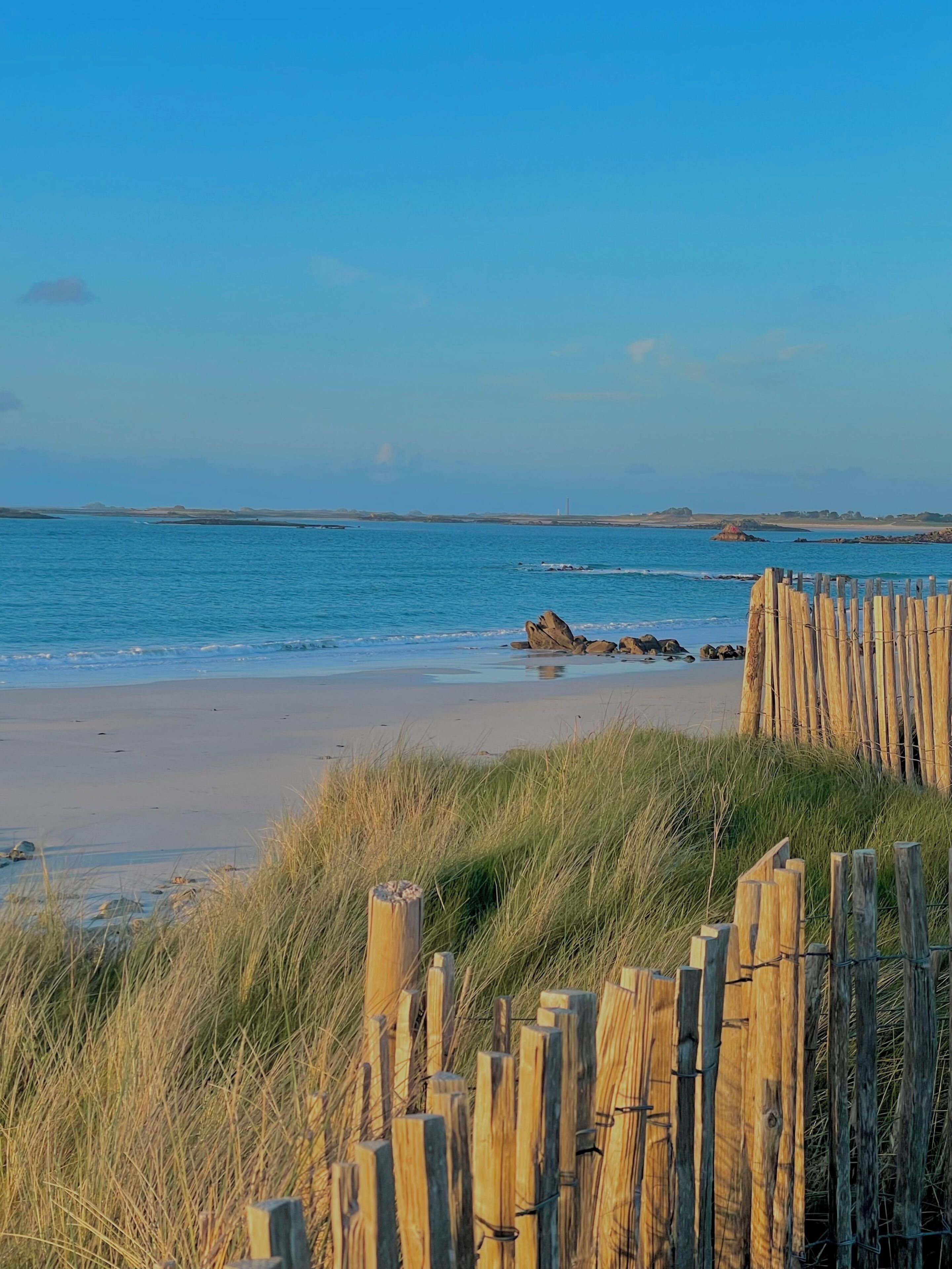 A beach with a wooden fence in the foreground, a sandy shore, and calm blue water.