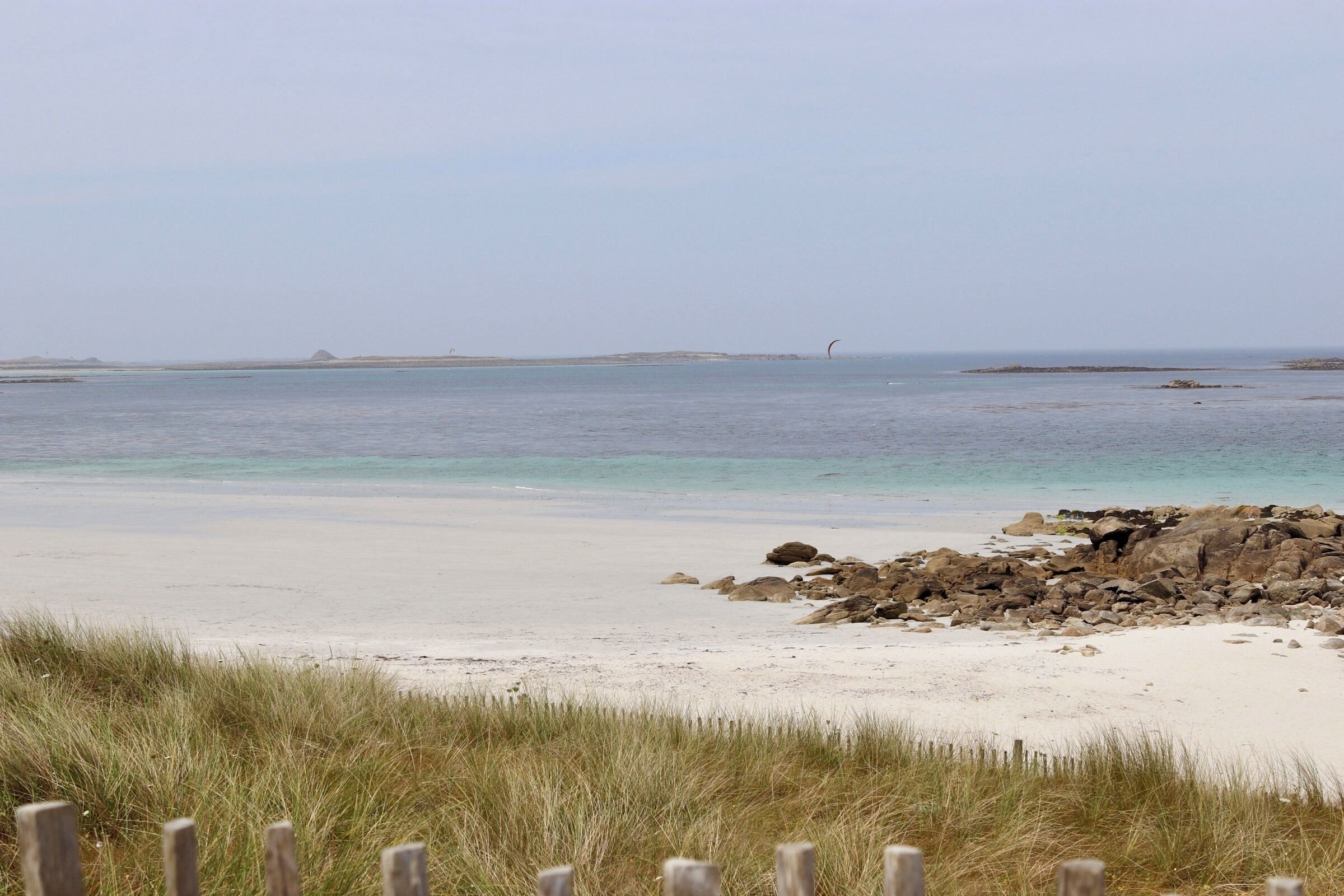 A beach with clear water, sandy shore, and a wooden fence in the foreground.