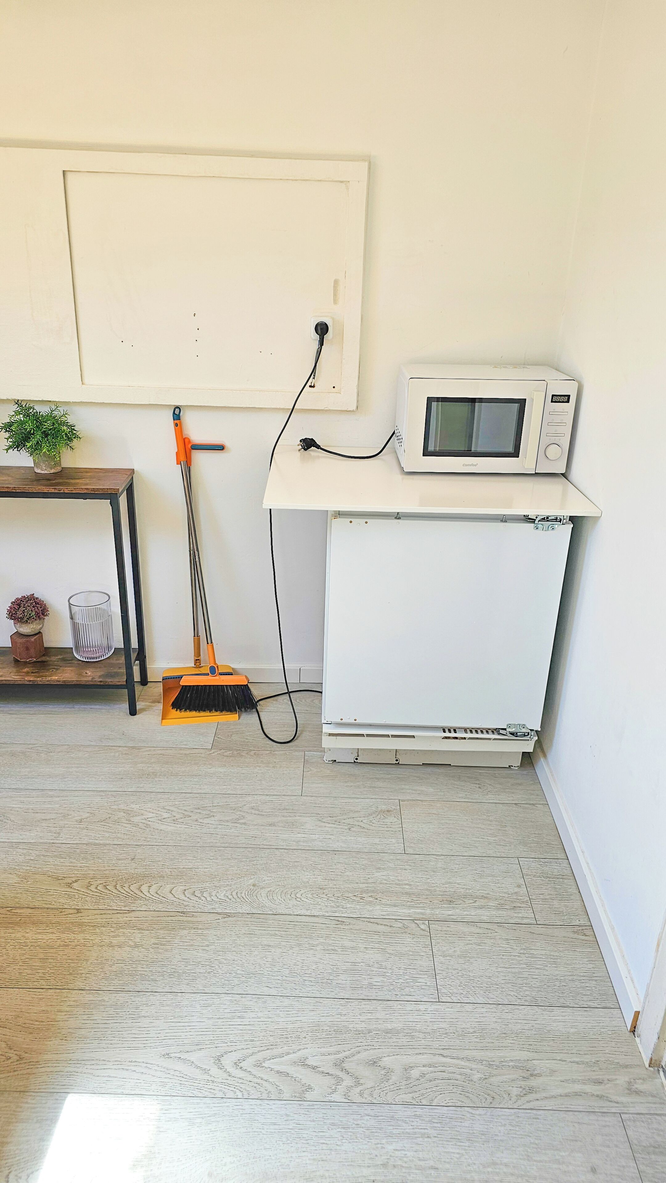 Una habitación con pared blanca, microondas blanco, escoba, toma de corriente y una pequeña mesa con una planta.