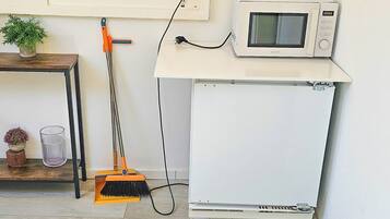 Una habitación con pared blanca, microondas blanco, escoba, toma de corriente y una pequeña mesa con una planta.