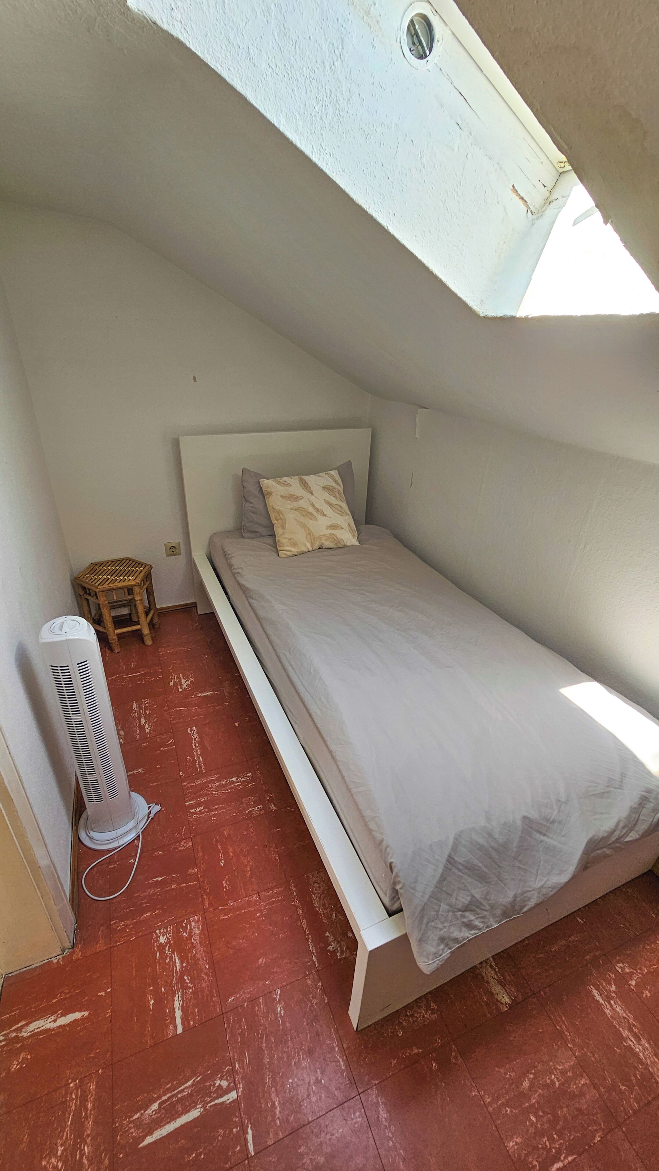 A small, single-bed room with a sloped ceiling, a standing heater, and a small stool.