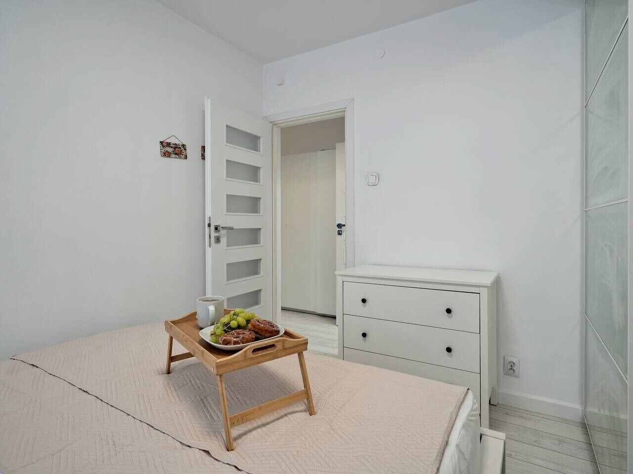 A bedroom with a bed, a bedside table with a tray of fruit, a white dresser, and a door leading to another room.