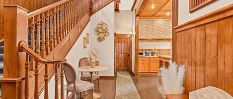 A wooden staircase leading to a dining area with a round table and chairs, a kitchen with wooden cabinets, and a fluffy white accent on a table.