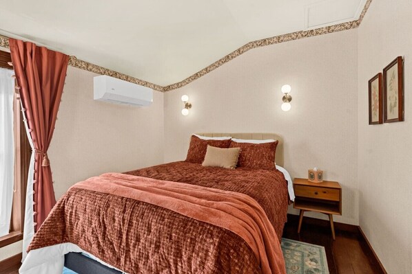Un dormitorio con una cama, una mesita de noche, aire acondicionado instalado en la pared y una ventana con cortinas.