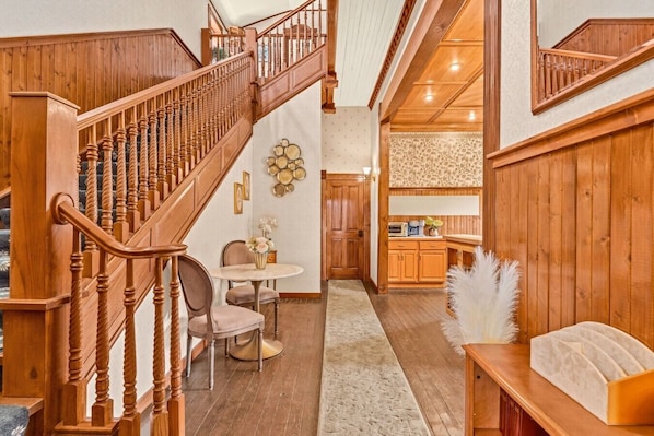 A wooden staircase leading to a dining area with a round table and chairs, a kitchen with wooden cabinets, and a fluffy white accent on a table.