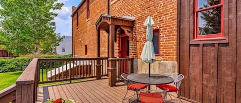 A wooden deck with a table and chairs, a red door, and a brick building in the background.