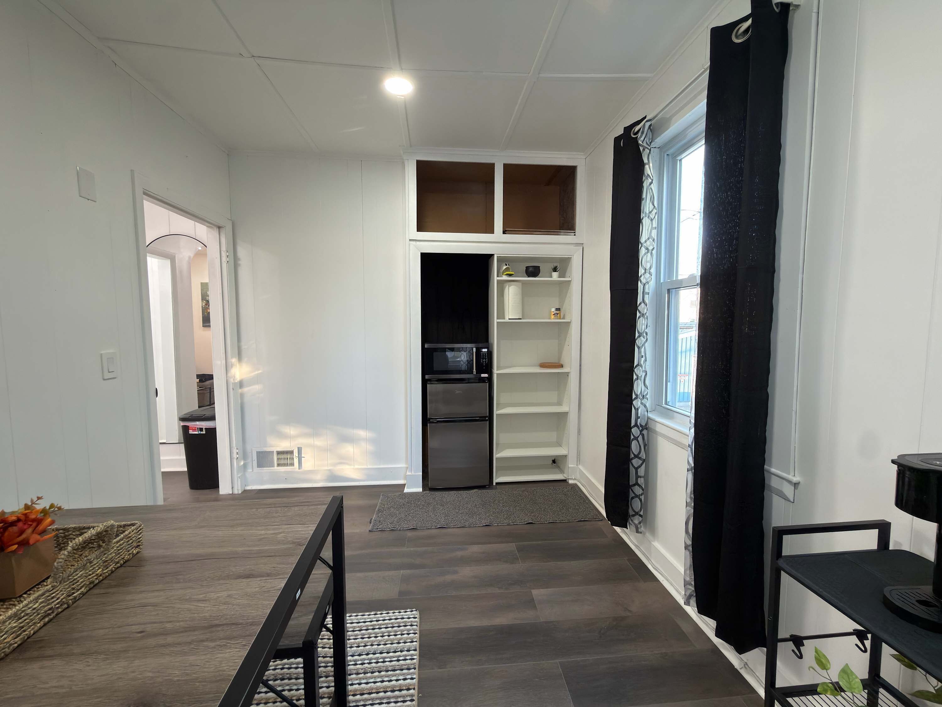 Una cocina moderna con armarios blancos, horno empotrado y microondas. Hay un comedor con una mesa, sillas, una alfombra y una ventana con cortinas negras.