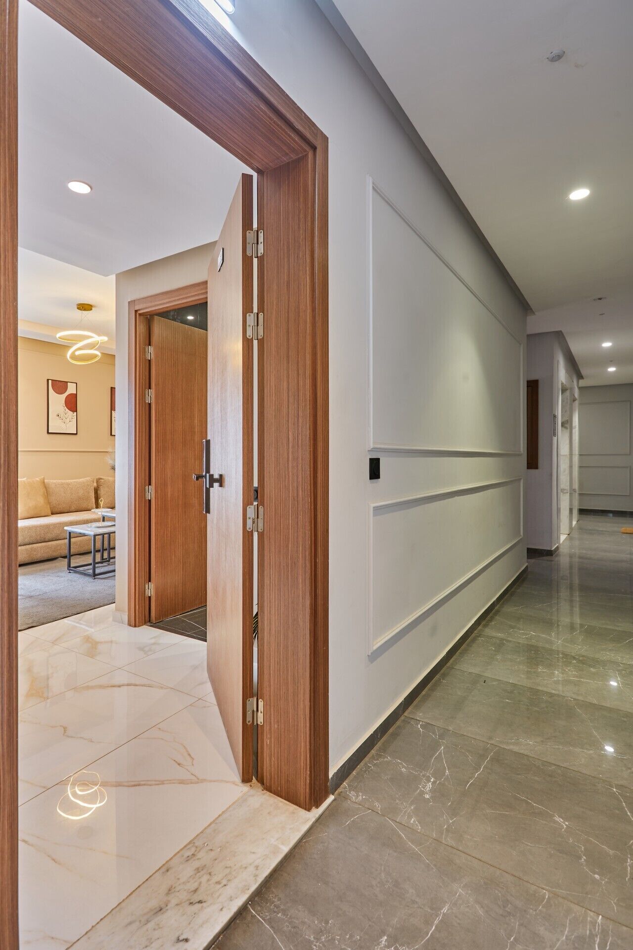 A hallway with a wooden door, a marble floor, and a white wall with a recessed light fixture.