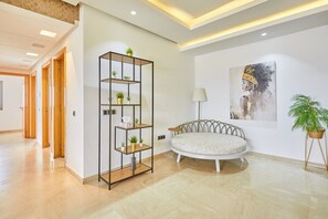 Een moderne woonkamer met een ronde witte bank, een hoge zwarte metalen boekenkast met planten, een vloerlamp en een schilderij aan de muur.