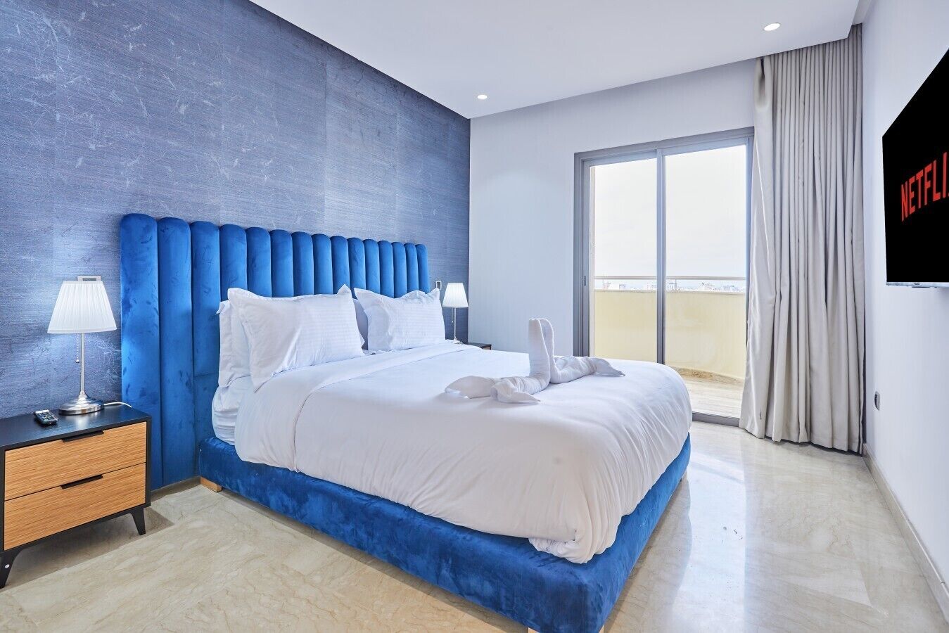 A modern hotel room with a large bed, a blue headboard, a TV on the wall, and a balcony view.