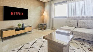 A modern living room with a flat-screen TV displaying 'NETFLIX,' a sofa, a coffee table, a side table with a lamp, and a large window with curtains. - Luxury 3BR | Prime Casablanca | Designer Stay (Casablanca)