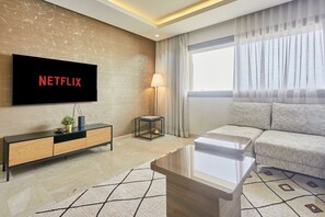 Ett modernt vardagsrum med en platt-TV som visar "NETFLIX", en soffa, ett soffbord, ett sidobord med en lampa och ett stort fönster med gardiner.