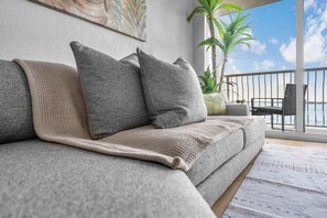 Ein graues Sofa mit einer beige farbenen Decke und Kissen, einer Topfpflanze und Blick auf das Meer durch ein Balkongeländer.