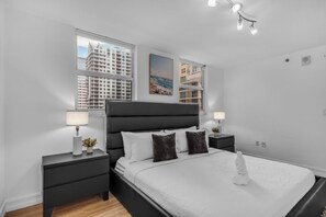 A modern bedroom with a large bed, two bedside tables, a window with a city view, and a ceiling light fixture. - Stunning Bay View 3BR | Pool, Gym & Brickell (Miami)