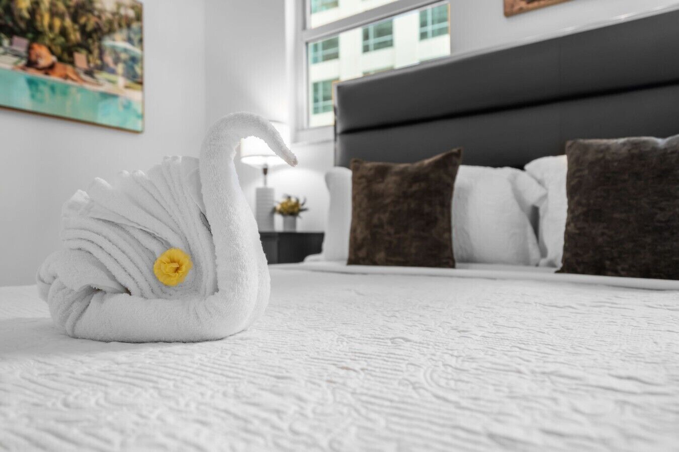 A swan-shaped towel on a bed with a white textured bedspread, a gray headboard, and a painting on the wall.