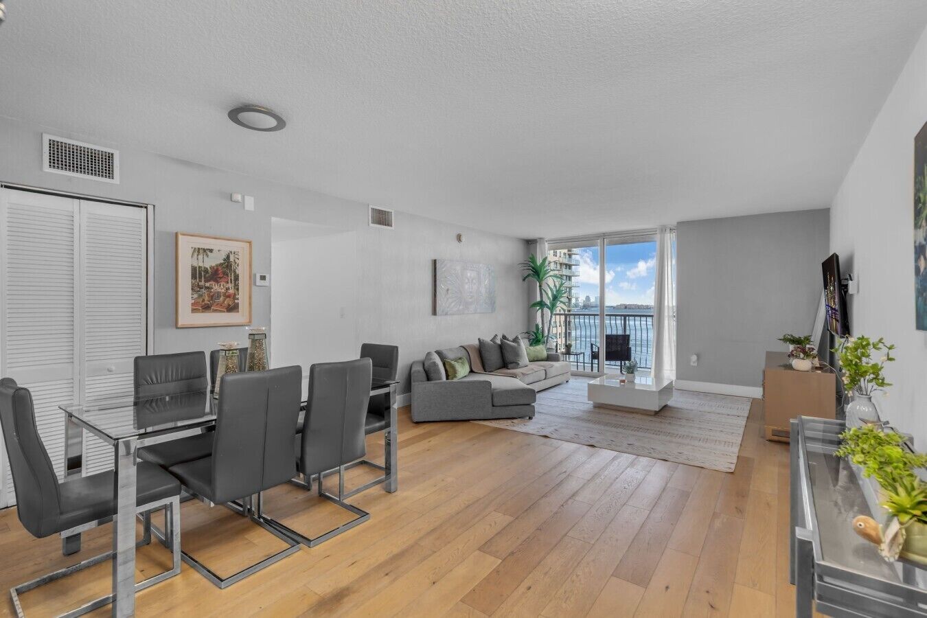 Una sala de estar moderna con comedor, un sofá gris, un televisor de pantalla plana y un balcón con vista al mar.