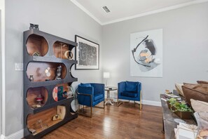 A modern living room with a dark wood shelving unit, blue armchairs, a round table, and a large abstract painting on the wall. - The Quincy - 4BR/2BA - Southwest Lubbock (Lubbock)