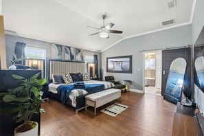 Ein Schlafzimmer mit einem Bett, einer Bank, einem Deckenventilator, einer Zimmerpflanze und Blick auf eine Küche.