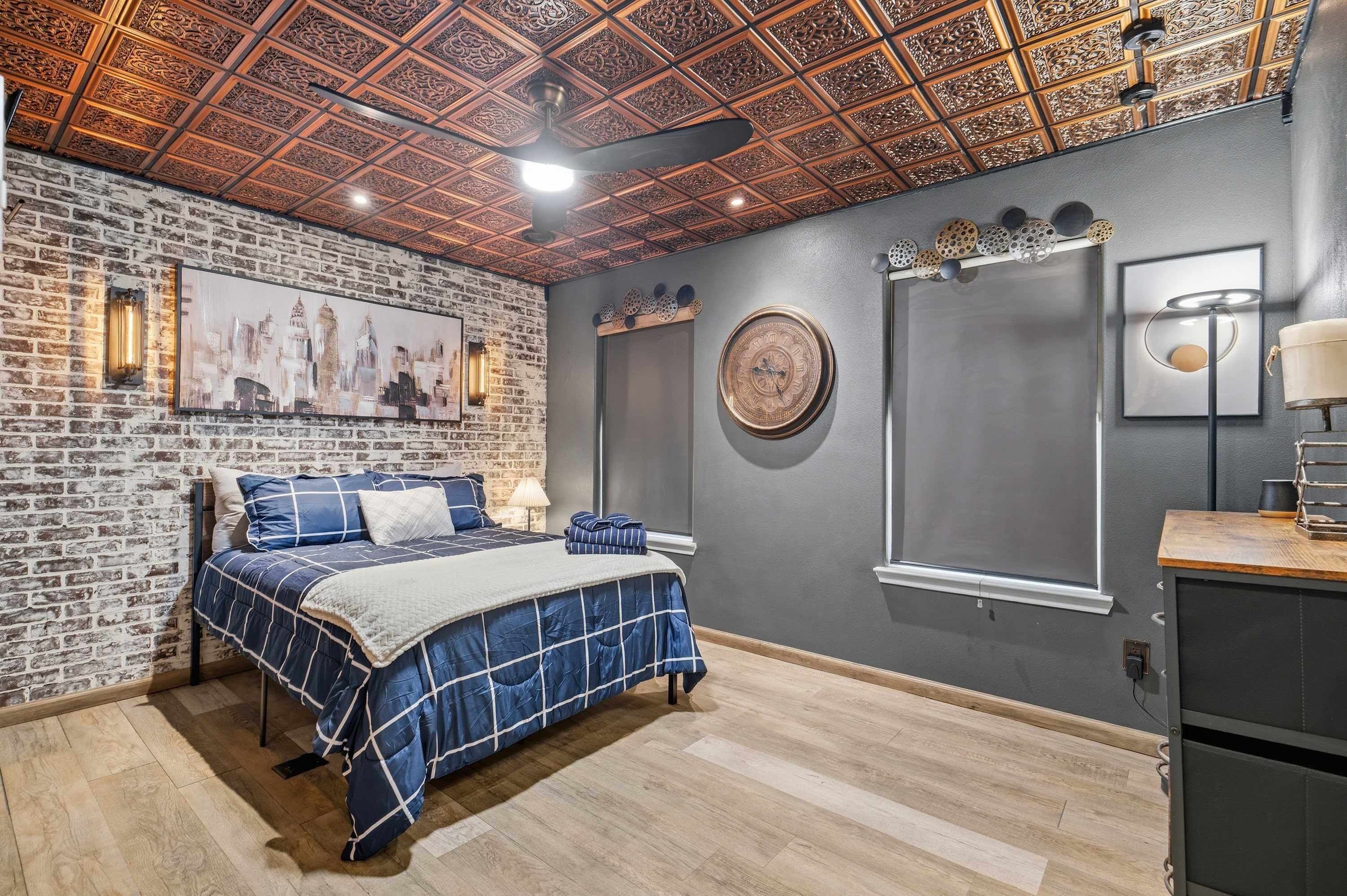 A bedroom with a brick wall, a bed with blue bedding, a decorative ceiling, and a cityscape painting.