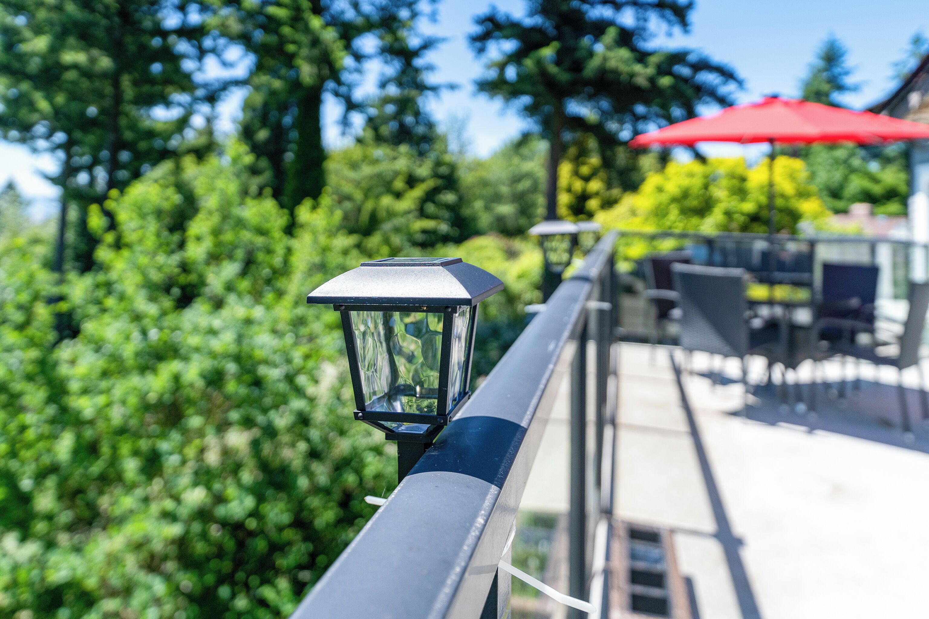 Une rambarde en métal avec un luminaire alimenté par énergie solaire, des meubles d’extérieur et un parasol rouge en arrière-plan.