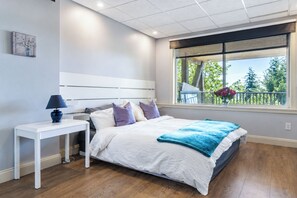 Et soveværelse med seng, sengebord, lampe og et vindue med udsigt til grønne omgivelser.