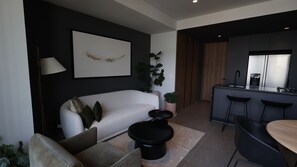 Ein modernes Wohnzimmer mit einer Couch, einem Couchtisch, einer Essecke mit Stühlen, einer Küche mit Kühlschrank und einer Zimmerpflanze.