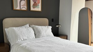 Una cama con cabecera color beige, ropa de cama blanca y dos cuadros abstractos en la pared.