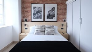 Un dormitorio con pared de ladrillo, una cama con ropa de cama blanca, dos cuadros enmarcados, una mesita de noche y un armario.