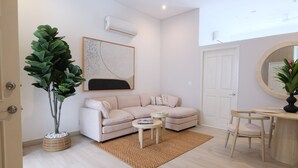 Una sala de estar con sofá, mesa de centro y sillas, una planta grande en maceta, un aire acondicionado de pared y un cuadro abstracto enmarcado.