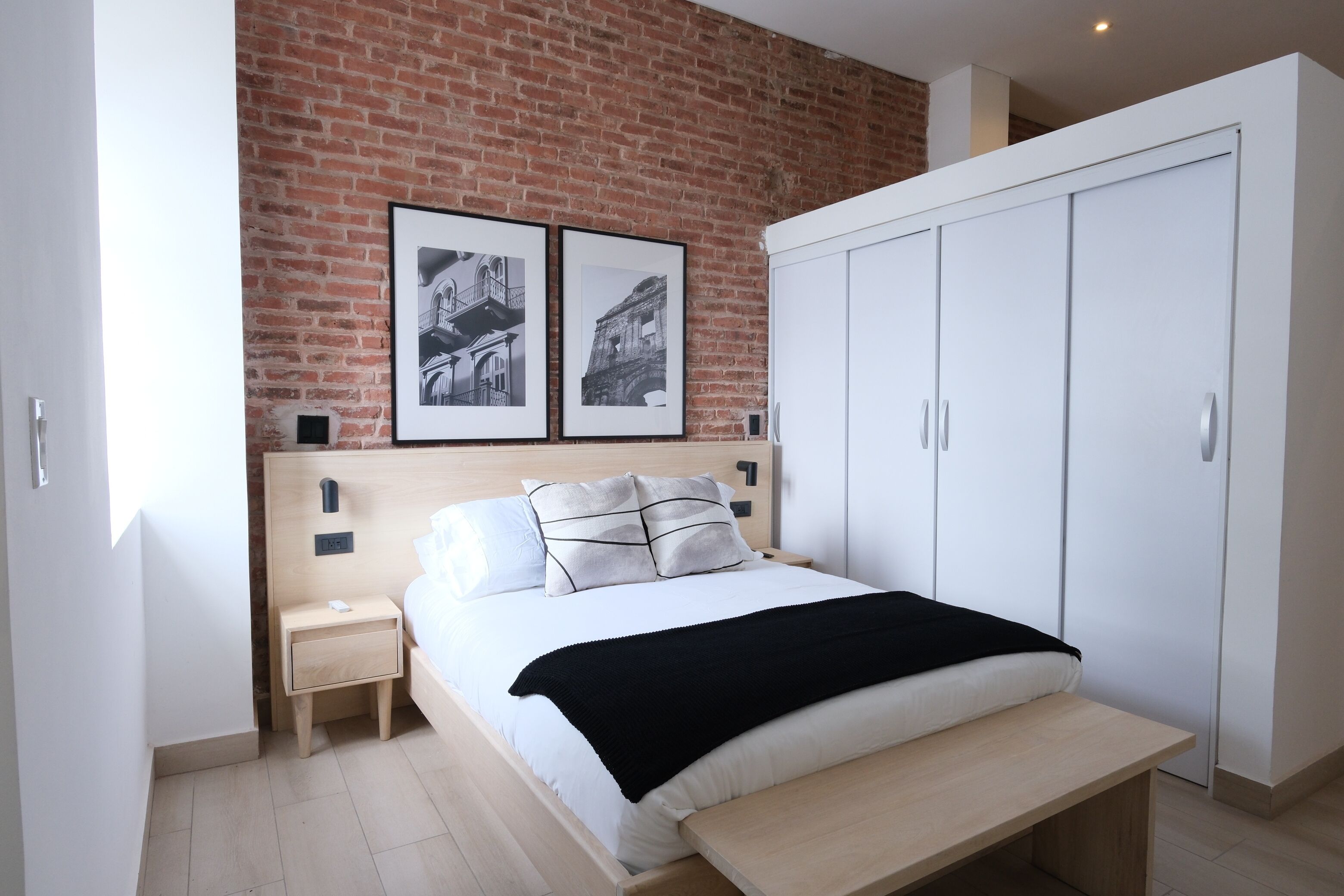 A bedroom with a brick wall, a bed with white bedding, two framed pictures, a white wardrobe, and a bedside table.