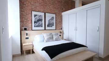 A bedroom with a brick wall, a bed with white bedding, two framed pictures, a white wardrobe, and a bedside table.