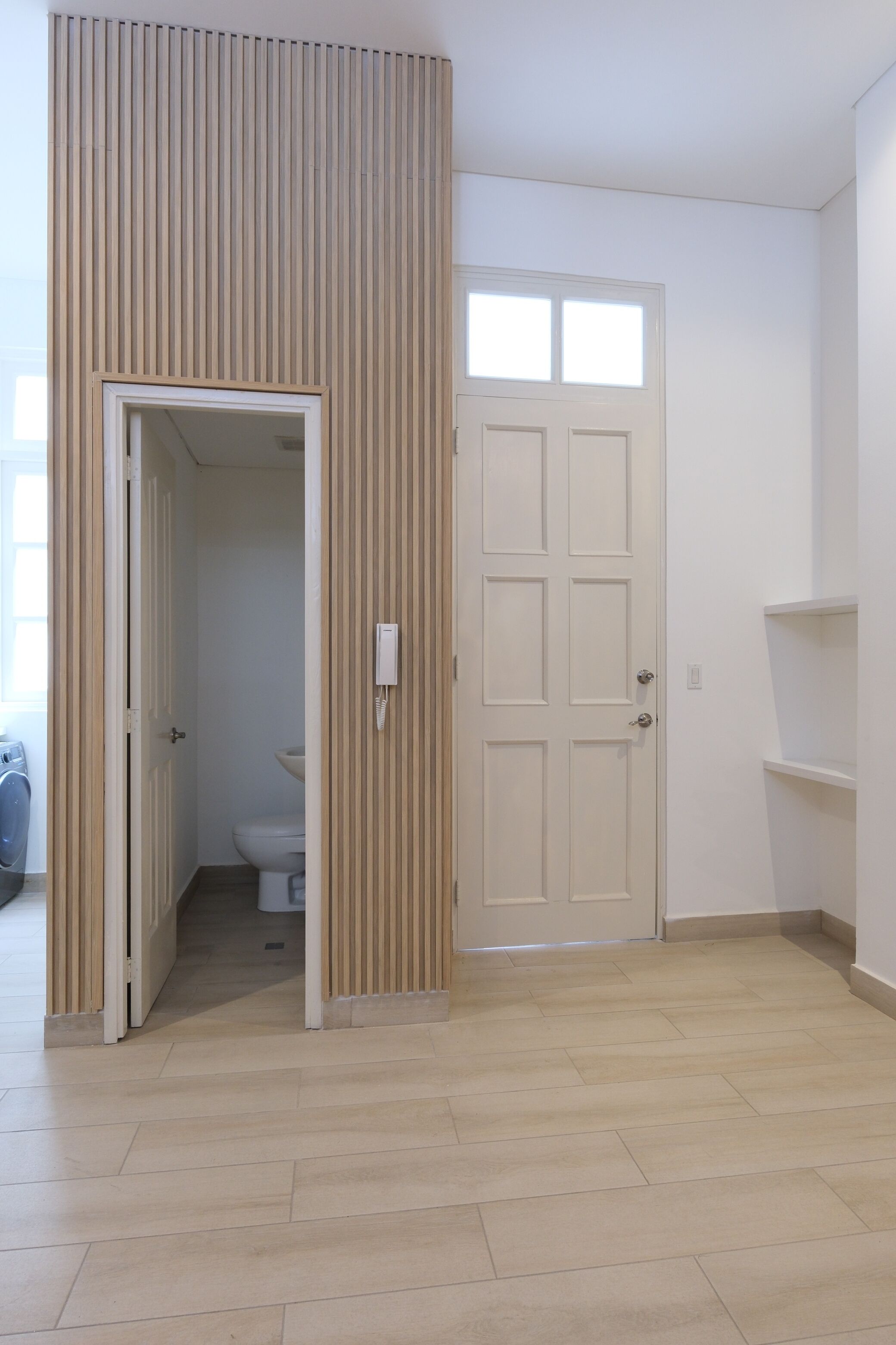 A room with a wooden slatted wall, a white door, a white toilet, and a light-colored floor.