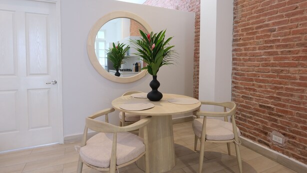 Un comedor con una mesa redonda, cuatro sillas, un florero con plantas, un espejo redondo y una pared de ladrillo.