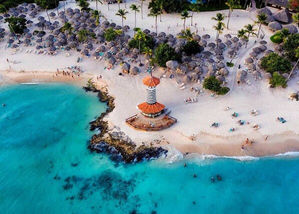 An aerial view of a beach with thatched umbrellas, a central structure with a red roof, and palm trees.