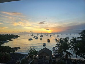 A coastal view with boats, palm trees, and thatched-roof structures.