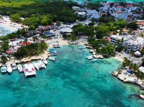 An aerial view of a coastal town with clear turquoise waters, boats docked at a marina, and buildings with red roofs.