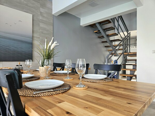 A dining area with a wooden table set for four, surrounded by chairs, with a staircase and a large painting on the wall.