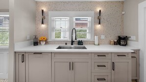 A kitchen with a white countertop, a stainless steel sink, and beige cabinetry. There are two wall-mounted lights and a window with blinds. - Mission Hills retreat, walkable, free parking (San Diego)