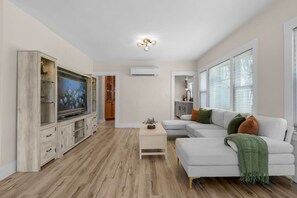 Ein modernes Wohnzimmer mit einem Ecksofa, einem Flachbildfernseher, Holzböden und einer Deckenleuchte.