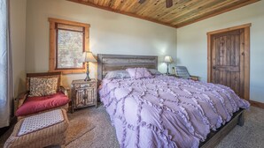 A bedroom with a wooden ceiling, a bed with a ruffled blanket, a wicker chair, a nightstand with a lamp, and a door. - Ridgeview Haven-perfect retreat (Fairplay)