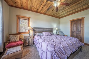 A bedroom with a wooden ceiling, a bed with a ruffled blanket, a wicker chair, a nightstand with a lamp, and a door. - Ridgeview Haven-perfect retreat (Fairplay)