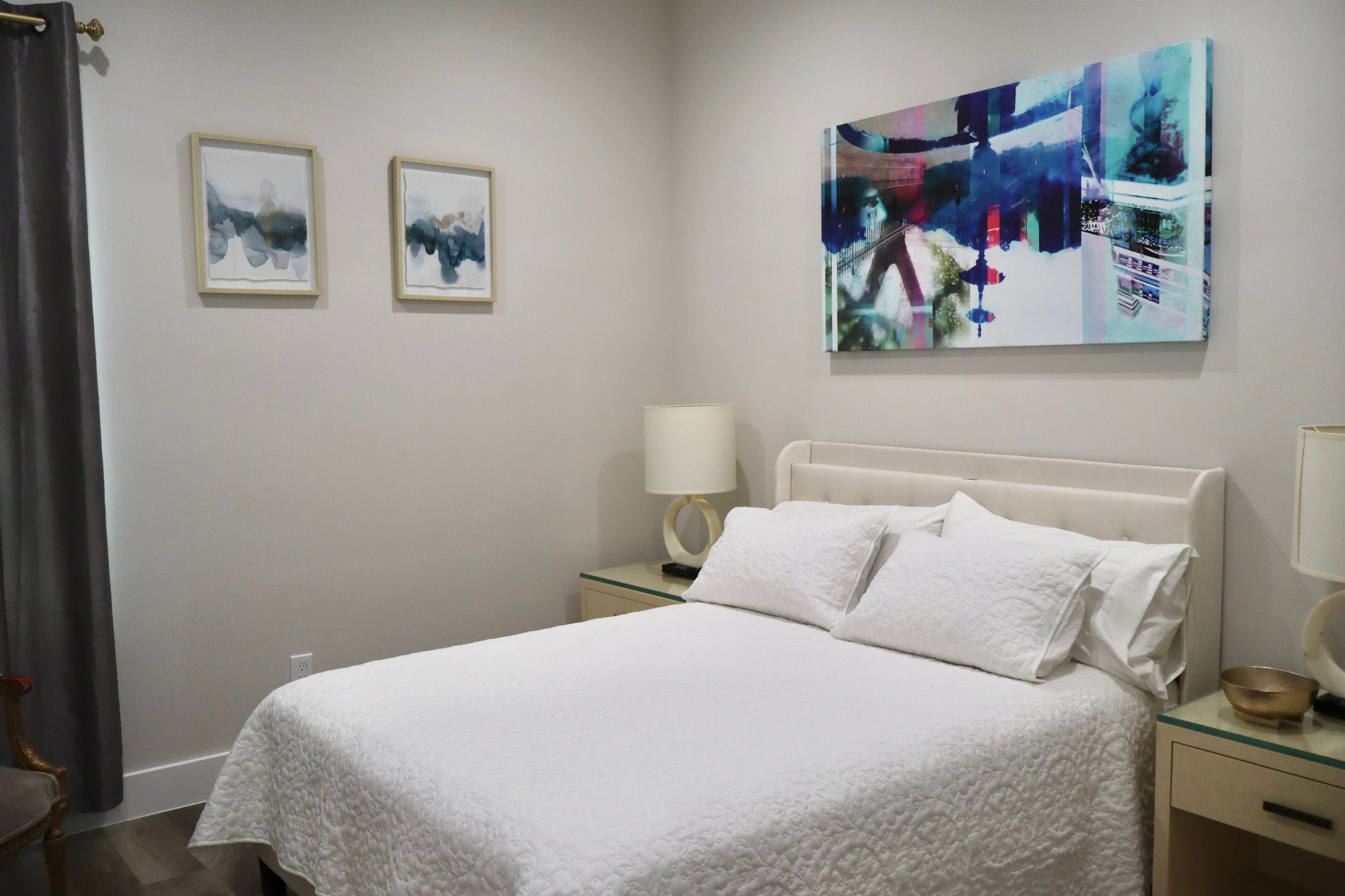 A hotel room with a bed, two bedside tables, a nightstand, a lamp, and two framed artworks on the wall.