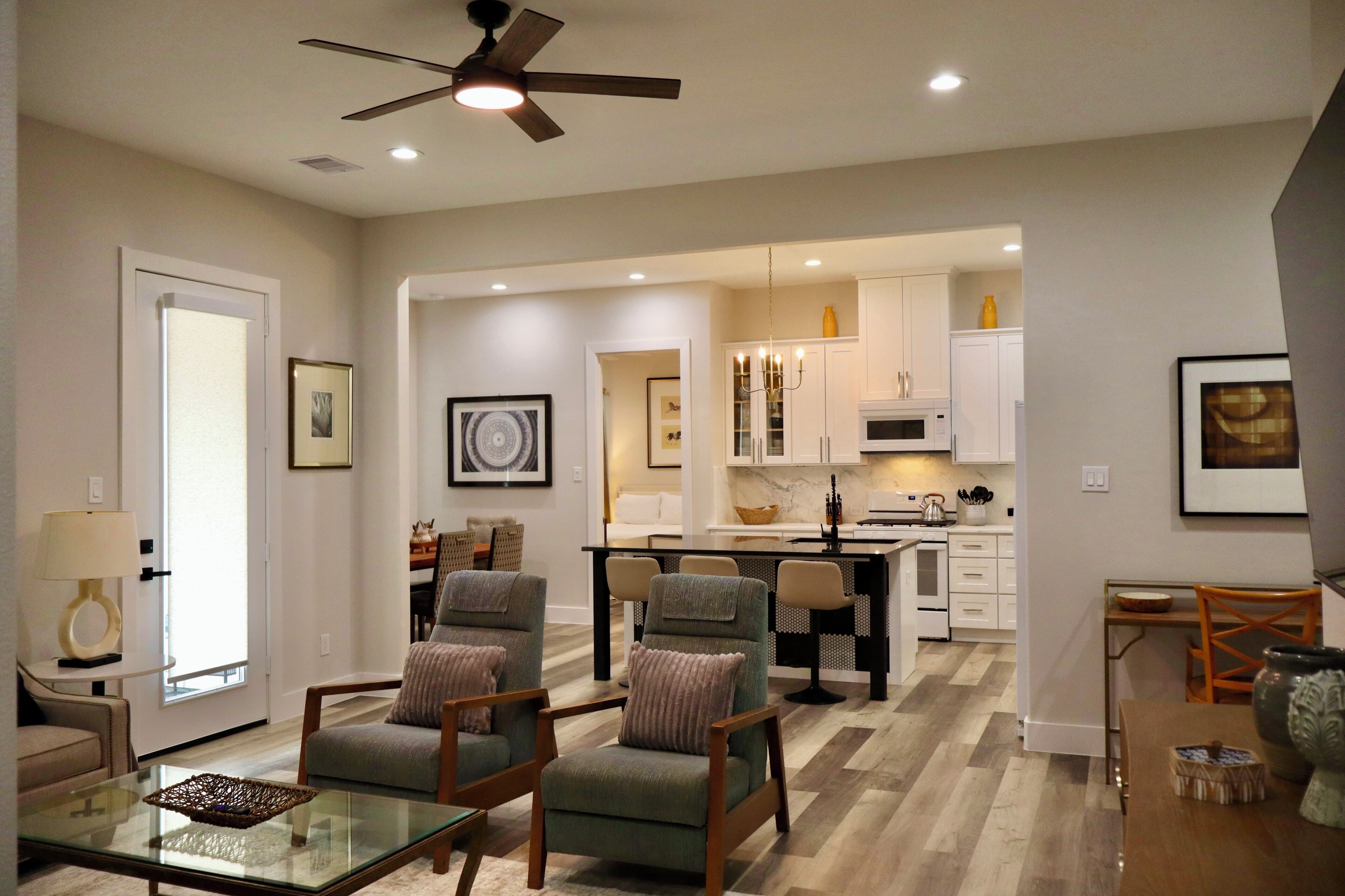 A modern living room with a glass coffee table, two armchairs, a ceiling fan, and a kitchen area with white cabinets and a microwave.